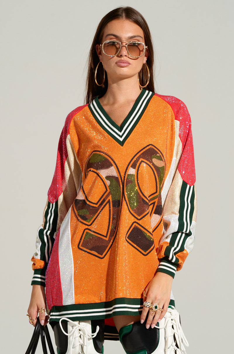 A woman with long brown hair, oversized sunglasses, and large hoop earrings wears the RYAN COLORBLOCK SEQUIN JERSEY MINI DRESS in orange, green, and red with the number 99 on it. The dress is adorned with sequins and has striped sleeves. She holds a small bag in one hand and stands against a grey background.