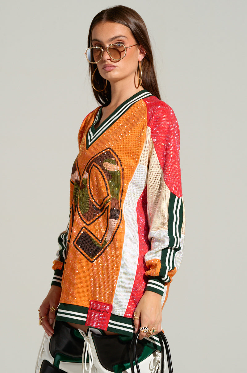 A person wearing an oversized, vibrant "RYAN COLORBLOCK SEQUIN JERSEY MINI DRESS" with the number "99" and a hint of camo print stands against a plain background. They have long hair, sunglasses, and hoop earrings. Their expression is neutral as they hold a strap or bag.