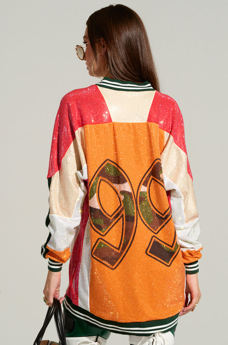 A woman with long brown hair stands facing away, wearing oversized sunglasses. She has on a vibrant Ryan Colorblock Sequin Jersey Mini Dress featuring red, white, orange, and green panels with a large number "99" on the back. She holds a black handbag in her left hand.