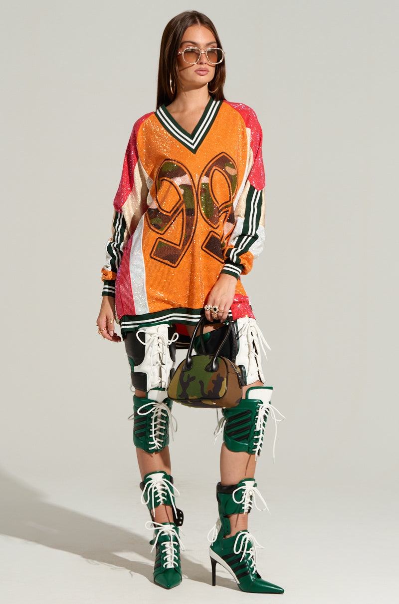 A woman stands confidently against a neutral background, wearing an oversized RYAN COLORBLOCK SEQUIN JERSEY MINI DRESS adorned with a vibrant camo print and the number 99. She pairs it with knee-high, lace-up boots and thigh straps, accessorizing with large sunglasses and carrying a small handbag.