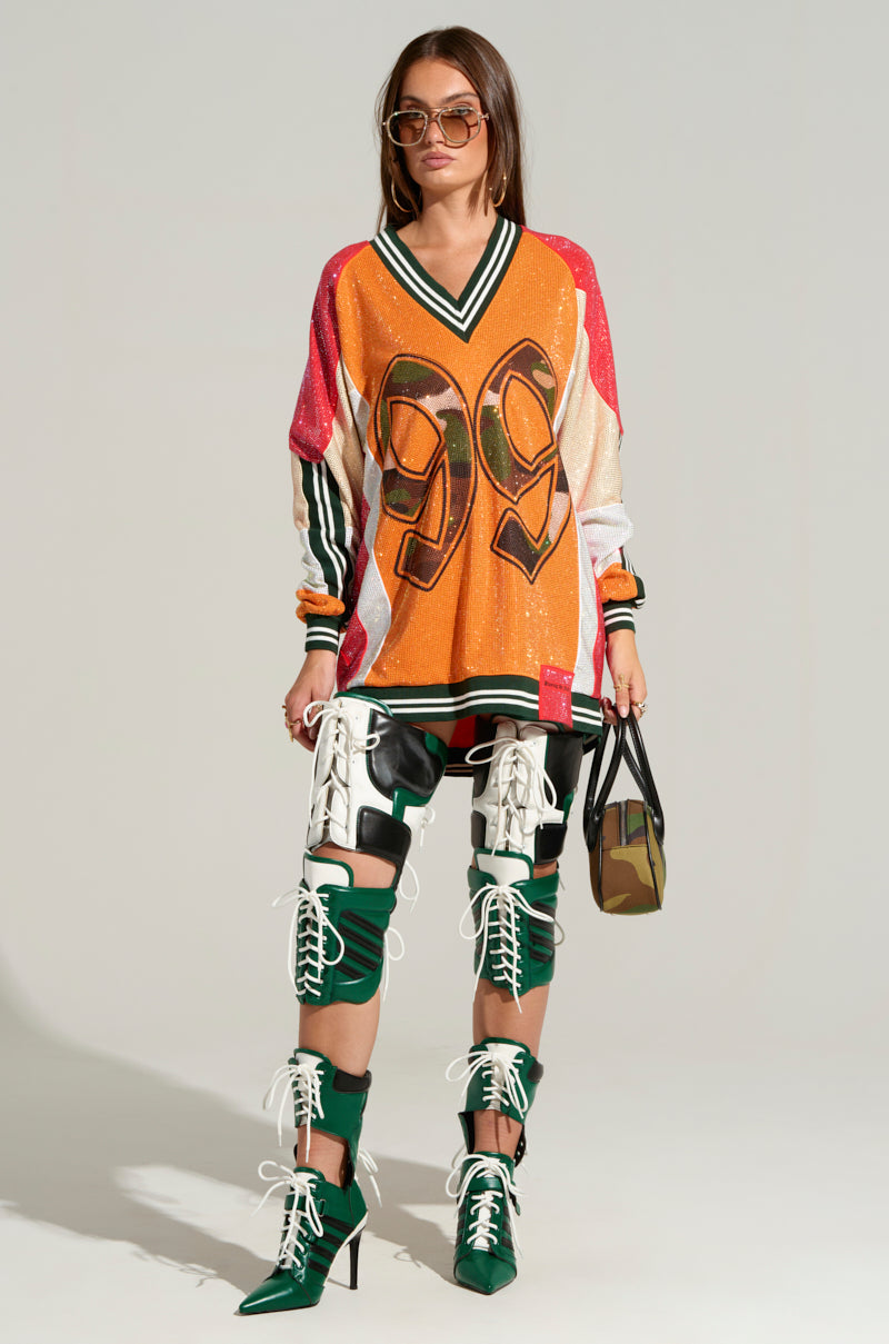 A person wearing an oversized orange sports jersey with the number 99, sunglasses, and carrying a RYAN COLORBLOCK SEQUIN JERSEY MINI DRESS in camo print. They have on green and white high-heeled lace-up boots with matching knee braces. The background is plain grey.
