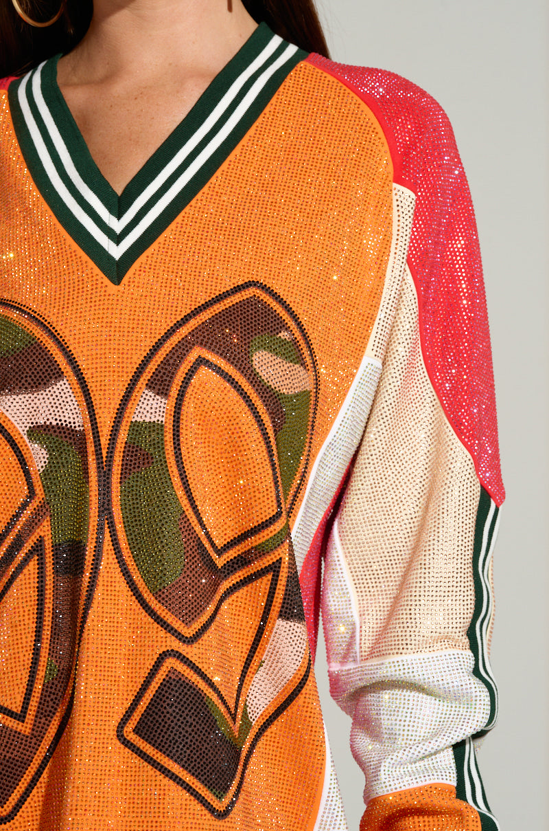 A person wearing the RYAN COLORBLOCK SEQUIN JERSEY MINI DRESS, featuring a vibrant mix of camouflage patterns and colors such as orange, red, and green with the number "99." The dress is designed with a V-neck collar accented by green and white stripes.