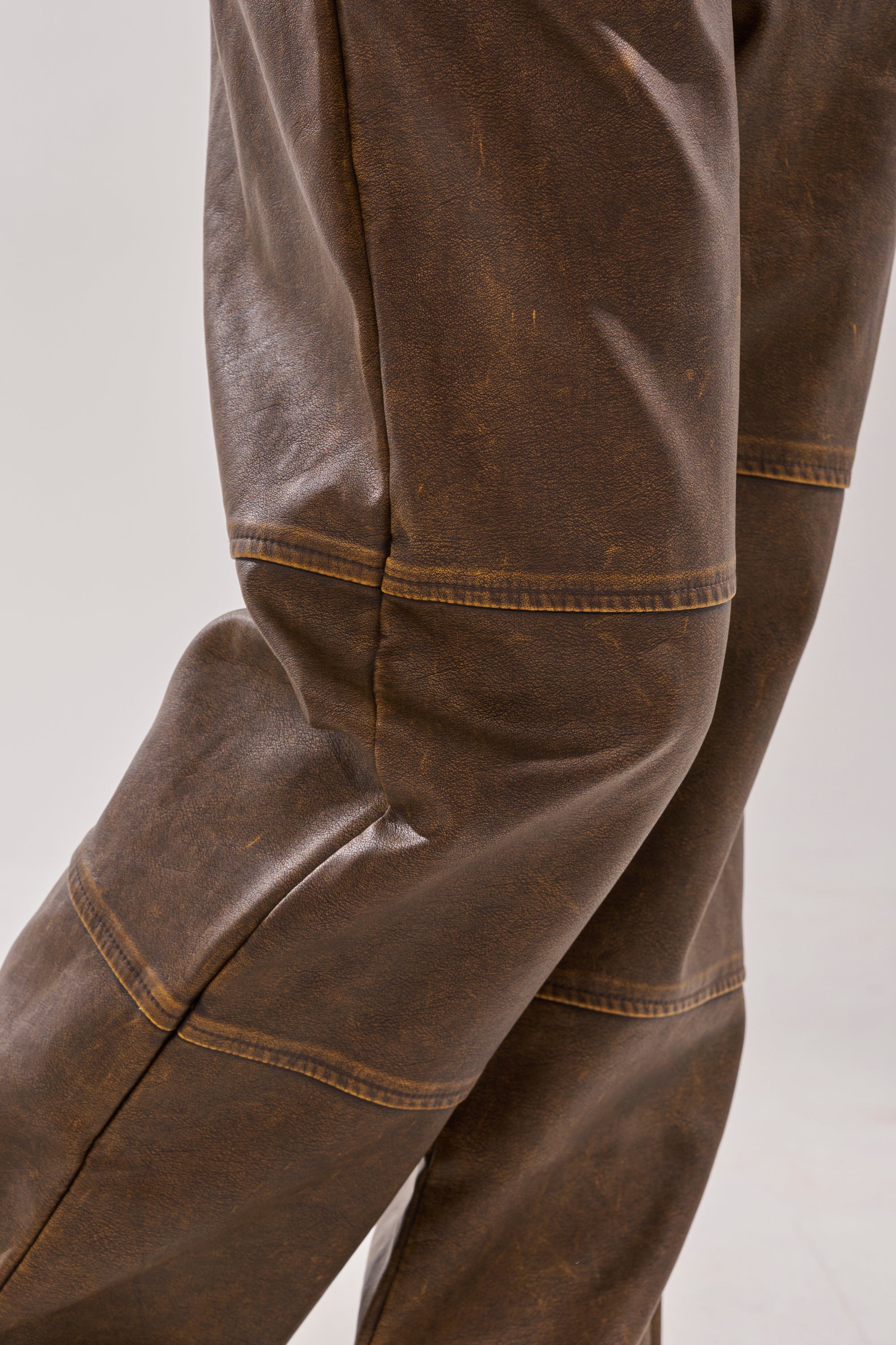 Close-up of a person wearing the ALL AROUND THE CITY TROUSER in brown faux leather with visible seams, textured finish, detailed stitching, and a straight-leg silhouette. Neutral, blurred background. 9