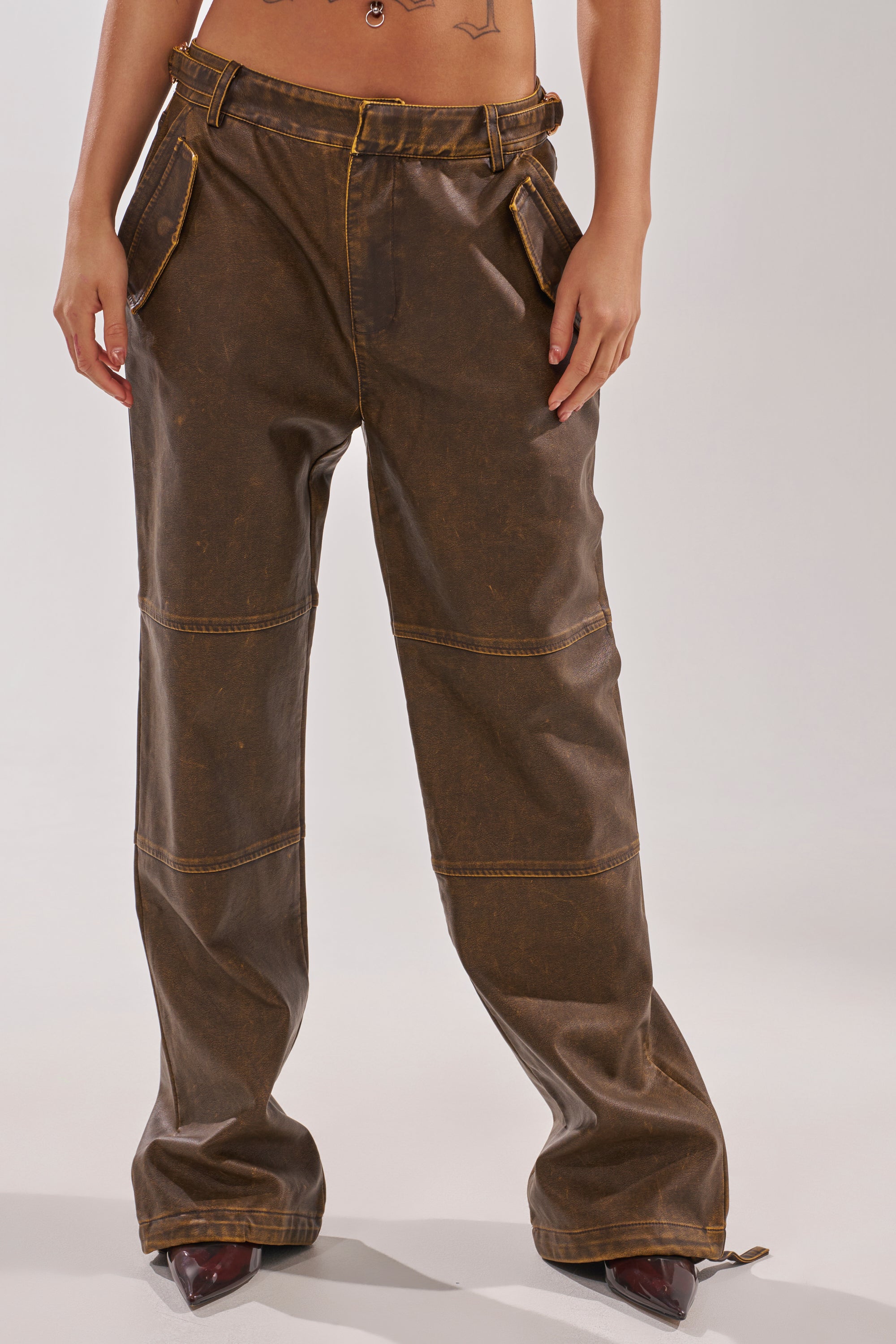 The lower torso and straight-leg silhouette of the ALL AROUND THE CITY TROUSER are shown; the brown, high-rise faux leather pants have visible stitching, front pockets, and a loose fit, with hands tucked into the pockets. 1