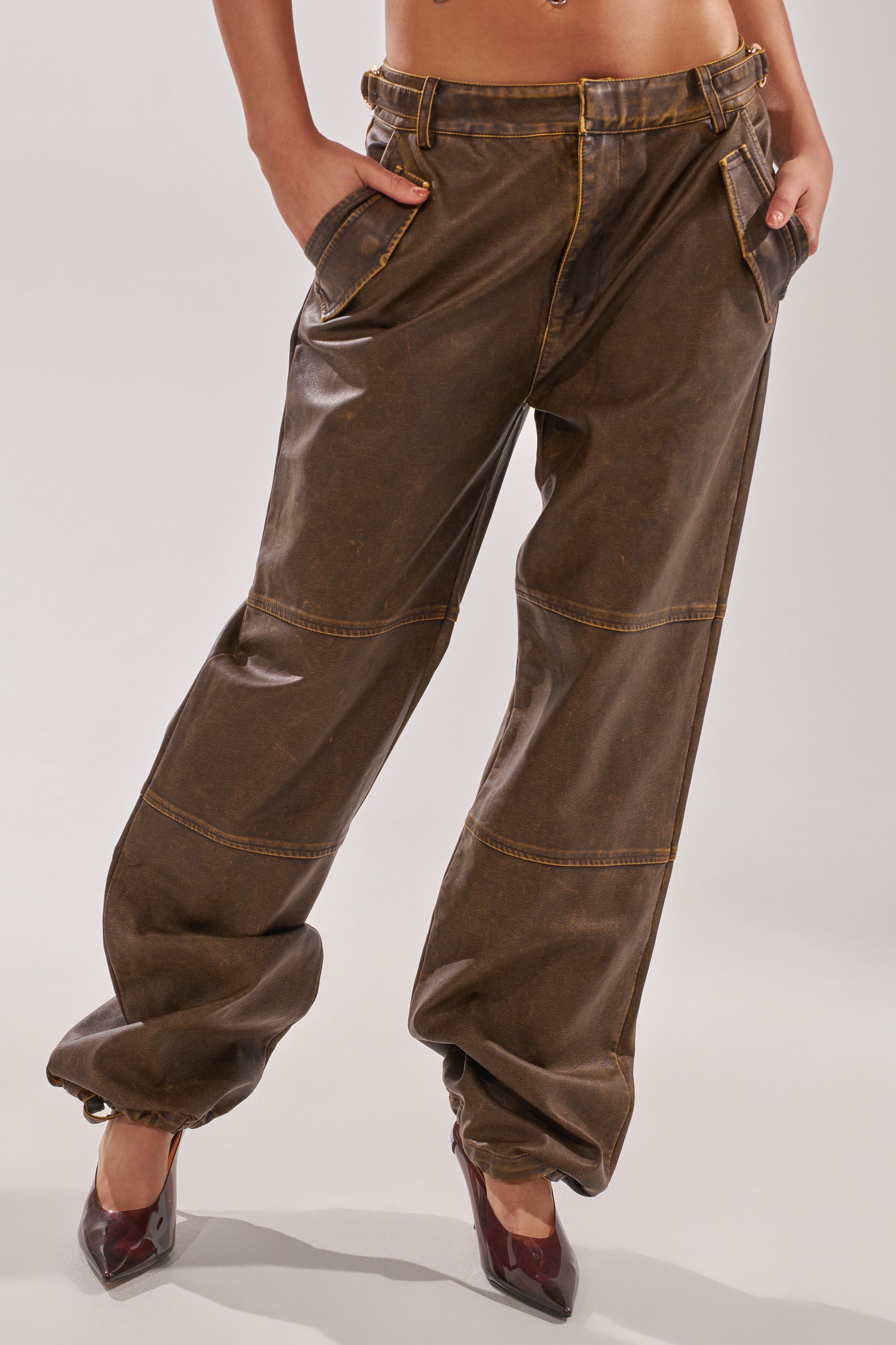A person models the ALL AROUND THE CITY TROUSER—high-rise, brown faux leather cargo pants with yellow stitching—paired with high-heeled burgundy shoes, hands in pockets, standing against a plain background. 2