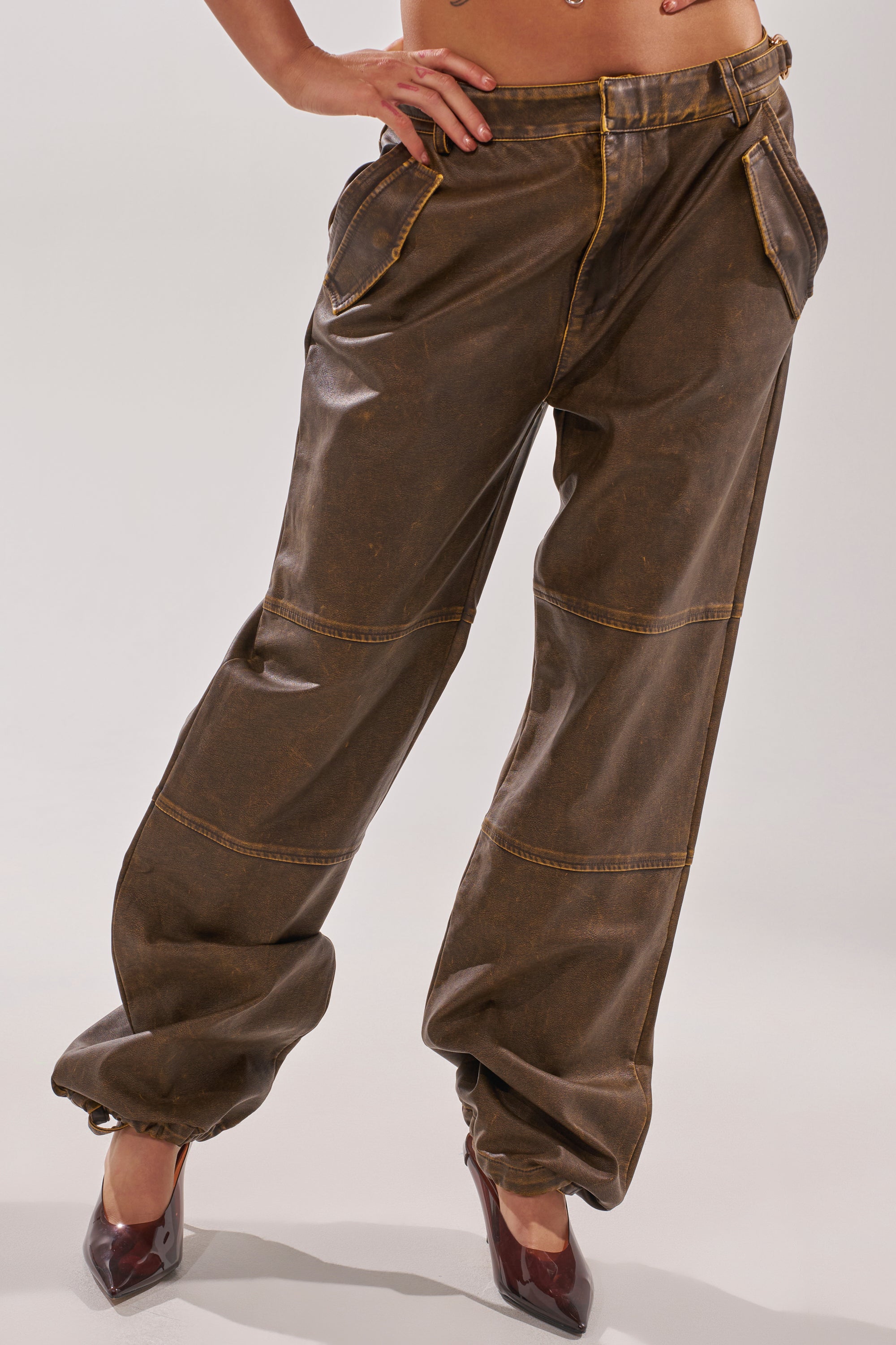 A person wears the ALL AROUND THE CITY TROUSER—loose, brown faux leather, high-rise, wide-leg with visible seams—paired with pointed burgundy heels; only their lower torso and legs are shown. 3