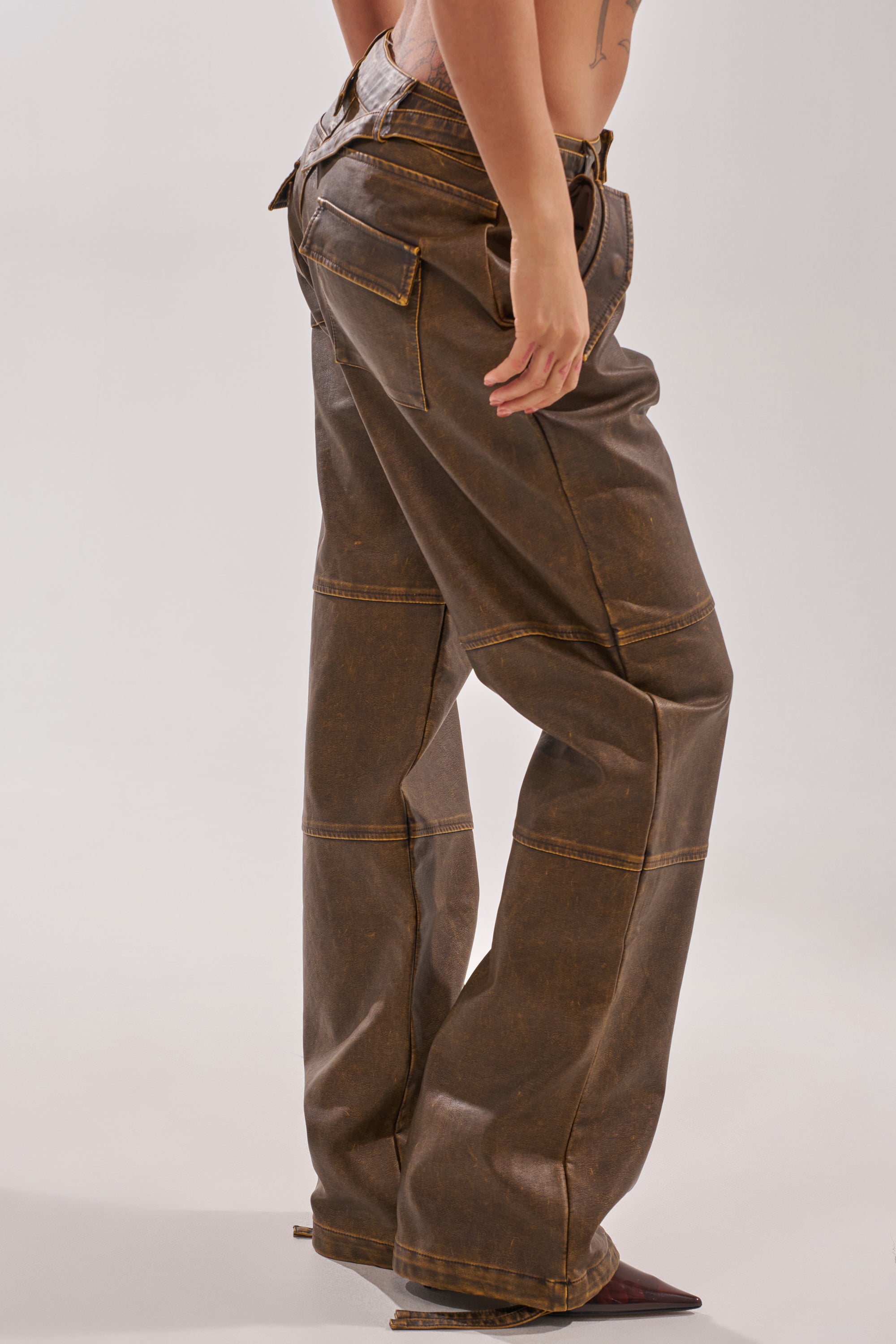A person models the ALL AROUND THE CITY TROUSER, featuring a high-rise fit, brown faux leather, visible stitching, and large back pockets, standing sideways against a plain background. 4