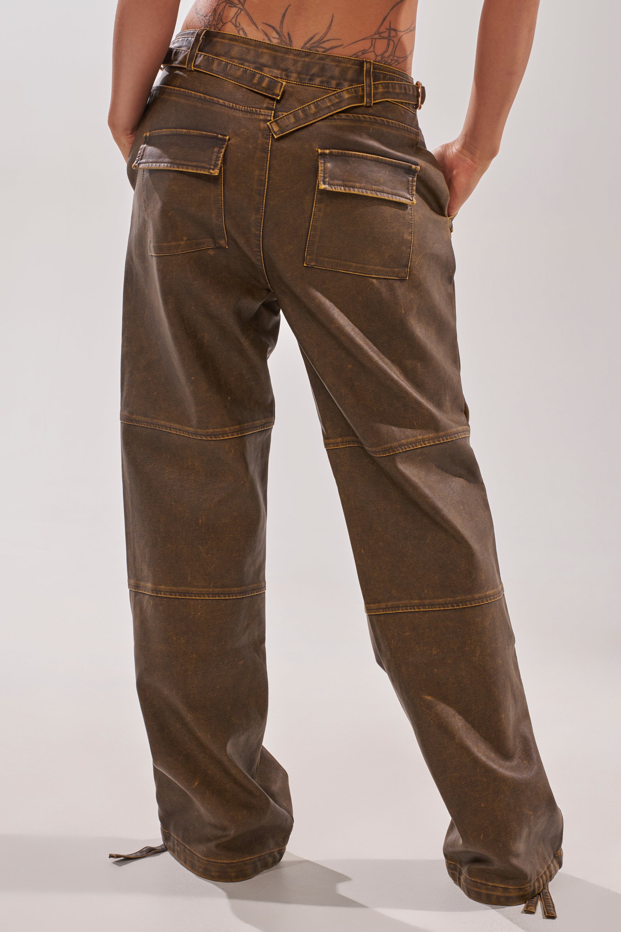 A person with a back tattoo wears the ALL AROUND THE CITY TROUSER—high-rise brown faux leather pants with a straight-leg fit, visible seams, flap back pockets, and adjustable ankle drawstrings—standing against a plain background. 5