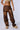 A person stands against a plain background, wearing a cropped top that reveals a "1999" tattoo on their stomach. They're sporting the OUT LIKE A BANDIT FAUX LEATHER CARGO PANT IN BROWN, featuring a high-rise, baggy silhouette with multiple pockets and zippers in a distressed finish, paired with fuzzy beige slippers. Their face is not visible.