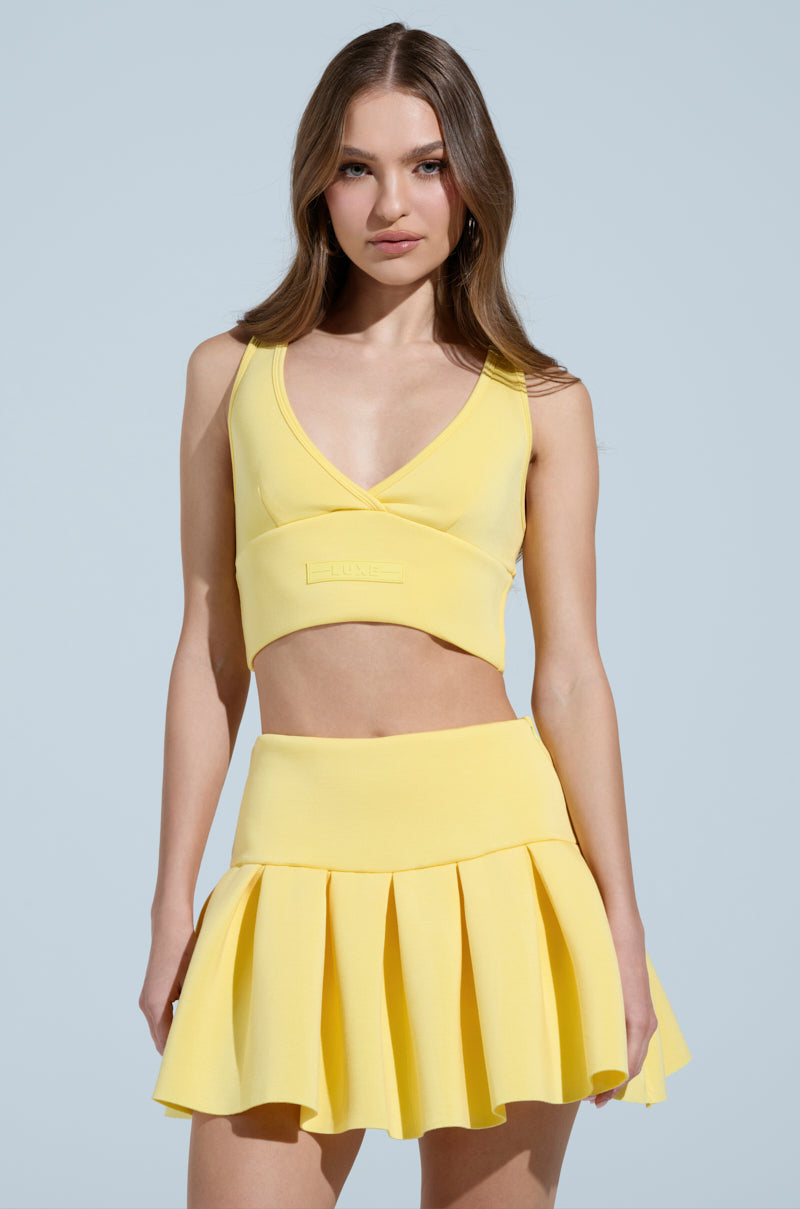 A woman radiates sporty-chic style in a yellow Paxton Superluxe Sleeveless Longline Bralette and matching pleated skirt, posing against a light blue backdrop. Her long brown hair flows freely as she confidently faces the camera, capturing effortless allure.