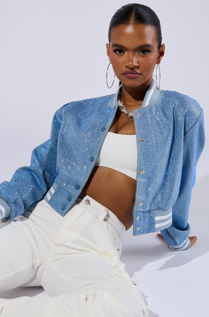 A person sits against a plain backdrop, wearing the HARLEY EMBELLISHED DENIM BOMBER with varsity-inspired stripe detailing, a white crop top, and high-waisted white pants. They have their hair pulled back and are accessorized with large hoop earrings and a statement necklace while looking confidently towards the camera. 0