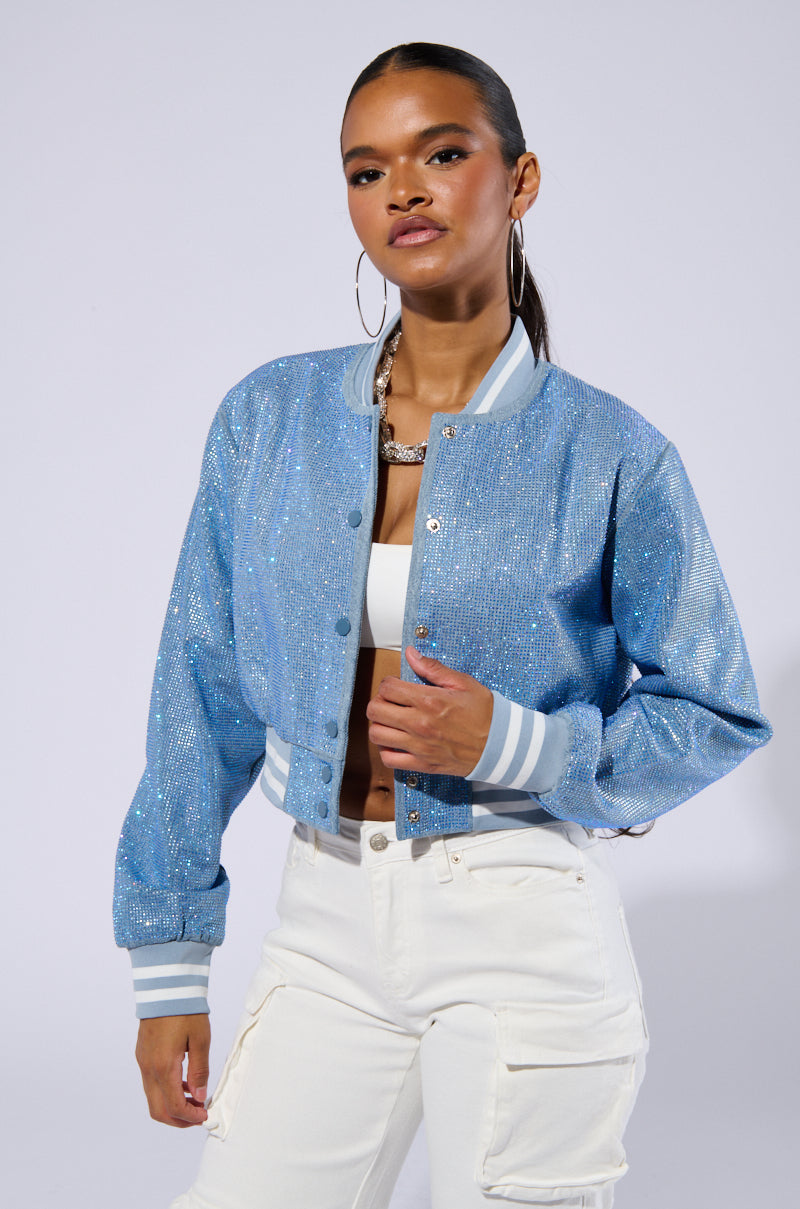 A person with long hair, wearing large hoop earrings, a white crop top, and white cargo pants poses confidently. They are also wearing the Harley Embellished Denim Bomber jacket with varsity-inspired stripe detailing on the cuffs and hem, which is unbuttoned to reveal the crop top. 1