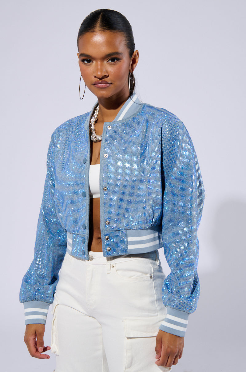 A woman stands against a plain backdrop wearing the HARLEY EMBELLISHED DENIM BOMBER over a white top and white pants. She has hoop earrings and a necklace. Her hair is tied back, and she gazes directly at the camera with a confident expression. 2