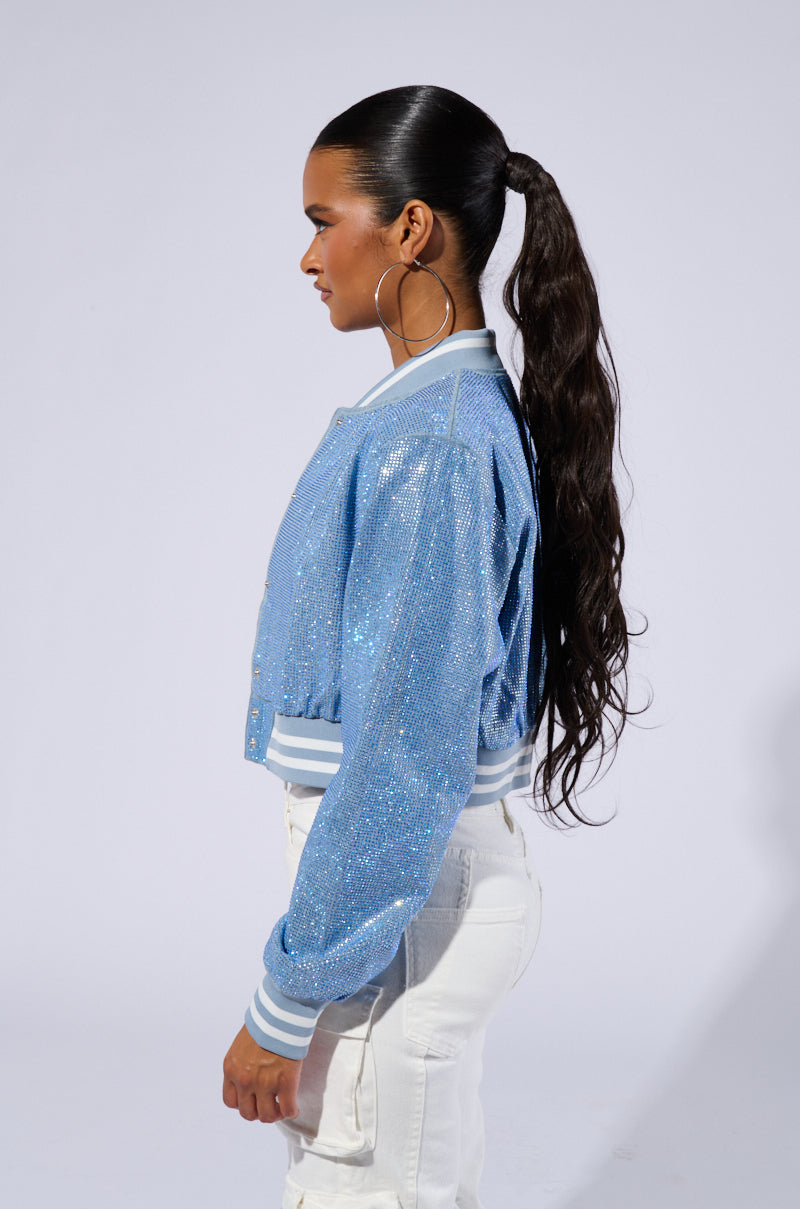 A person with long dark hair in a ponytail is shown in profile against a white background. They are wearing the HARLEY EMBELLISHED DENIM BOMBER, which features varsity-inspired stripe detailing, along with white pants. Large hoop earrings complete the outfit. 3