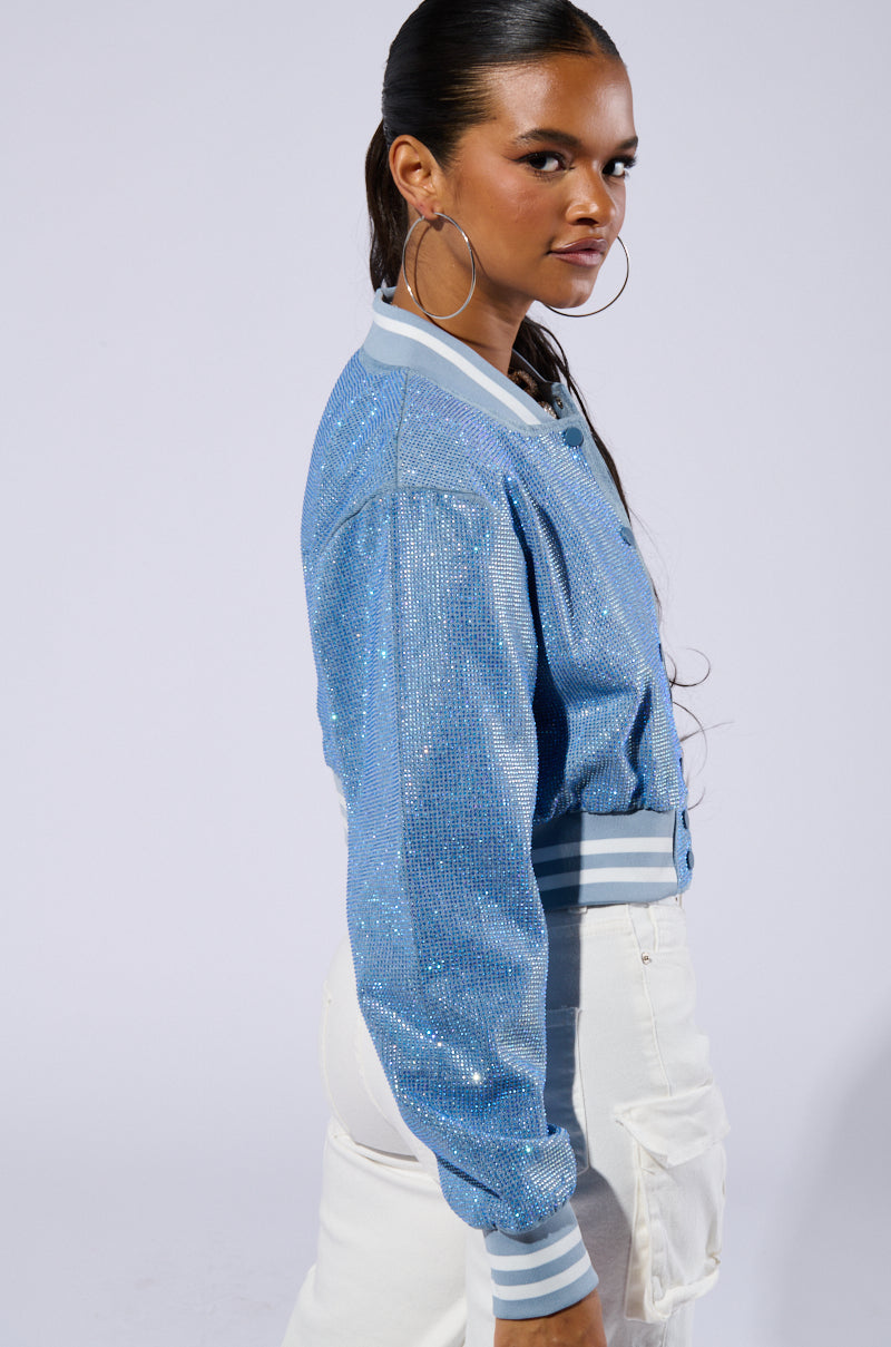 A woman with long, dark hair is facing sideways towards the camera. She is wearing large hoop earrings and the HARLEY EMBELLISHED DENIM BOMBER, which features a glittery light blue design and varsity-inspired stripe detailing on the cuffs and collar. She pairs it with white pants against a plain, light-colored background. 4