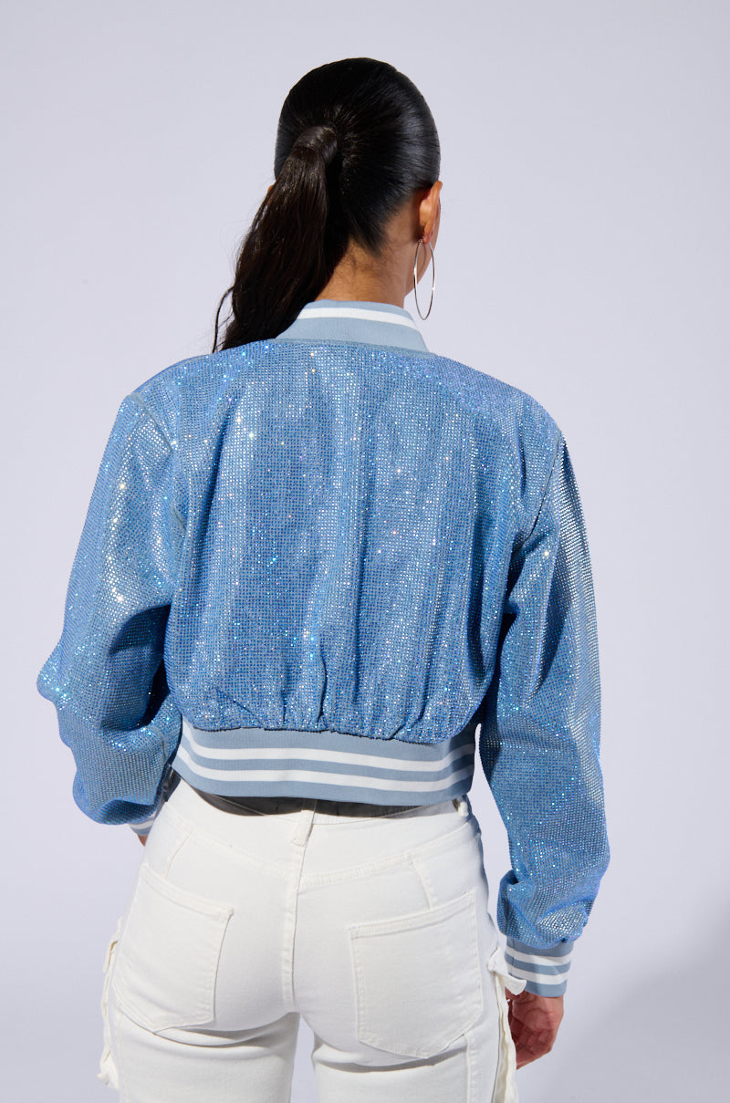 A person with long, dark hair in a ponytail is seen from behind wearing the HARLEY EMBELLISHED DENIM BOMBER, featuring varsity-inspired stripe detailing on the collar and waist. They are also wearing white pants with visible pockets on the sides. The background is plain light gray. 5