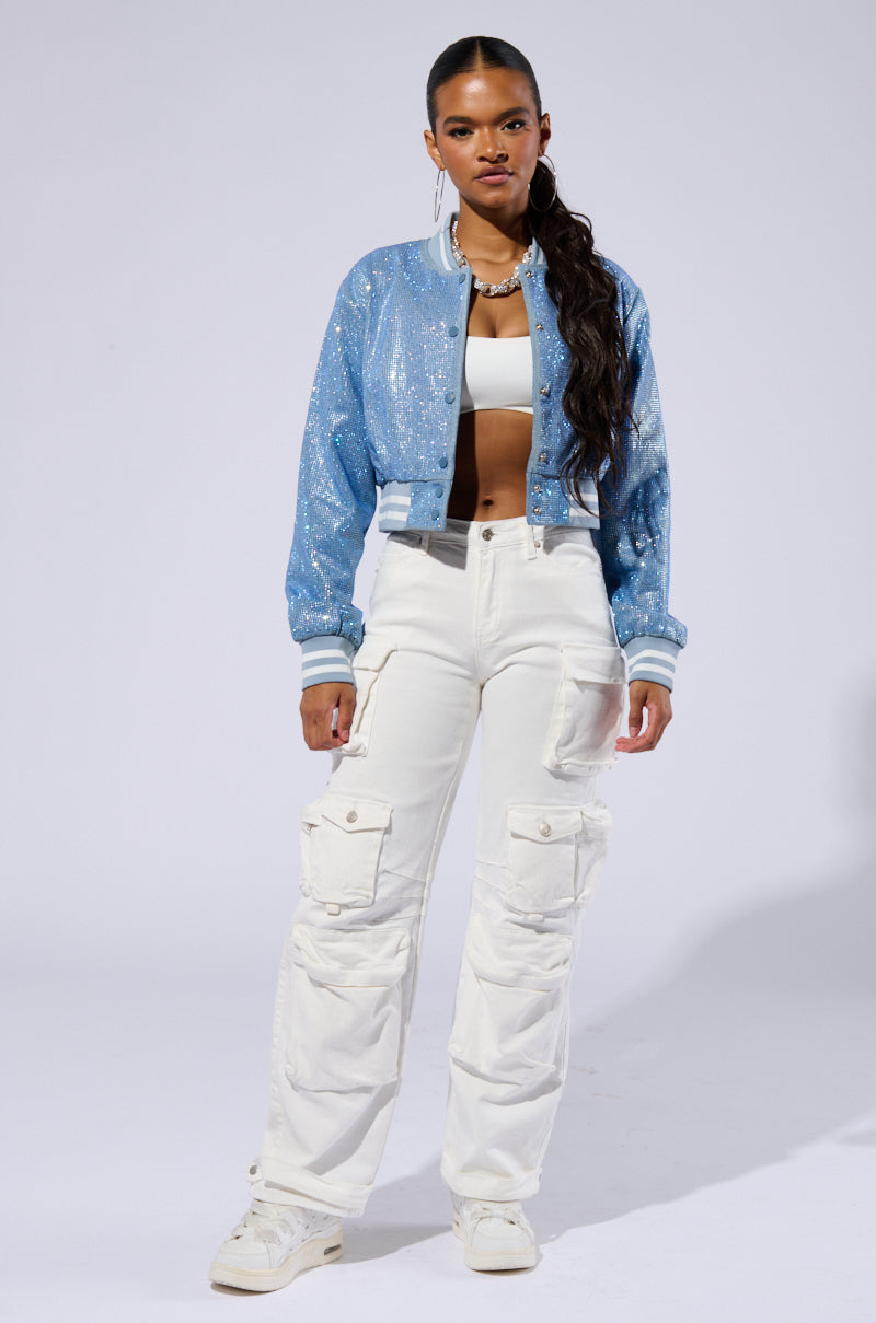 A woman stands confidently against a plain background. She wears the HARLEY EMBELLISHED DENIM BOMBER over a white crop top, paired with high-waisted white cargo pants and white sneakers. She has a long, sleek ponytail and accessorizes with a necklace and bracelets. 6