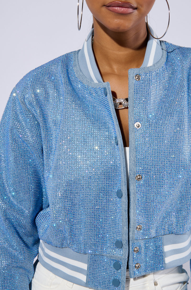 A person is wearing the HARLEY EMBELLISHED DENIM BOMBER with varsity-inspired stripe detailing on the collar and cuffs. Over a white top, the sparkly blue jacket features buttons and gives off a shimmering effect. The image captures only the person's lower face and upper torso. 7