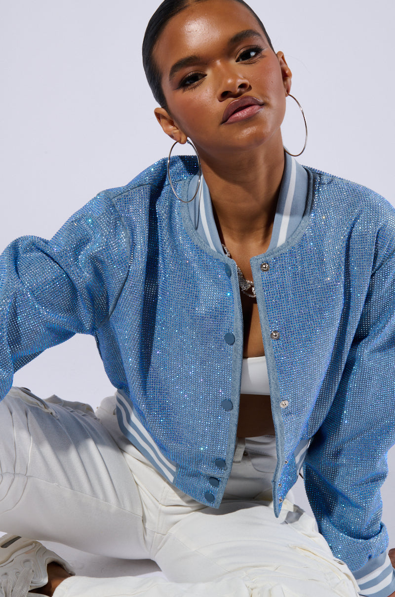 A person confidently poses in a HARLEY EMBELLISHED DENIM BOMBER, sparkling in vibrant blue and paired with large hoop earrings. They are also dressed in a white crop top and light-colored pants, with one knee on the ground. The plain white background accentuates the vivid blue of the jacket. 8