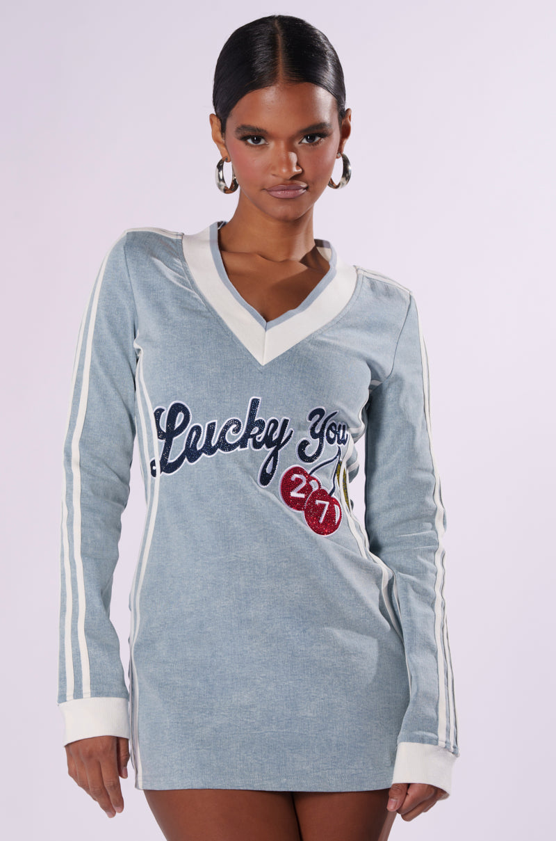 A person with dark hair pulled back wears the "With a Cherry on Top Denim Mini Dress," characterized by its light blue color, long sleeves, V-neck with a plunging neckline, and white accents. The dress also features the phrase "Lucky You 24/7" alongside a cherry graphic on the front. They stand against a plain light background, looking directly at the camera.