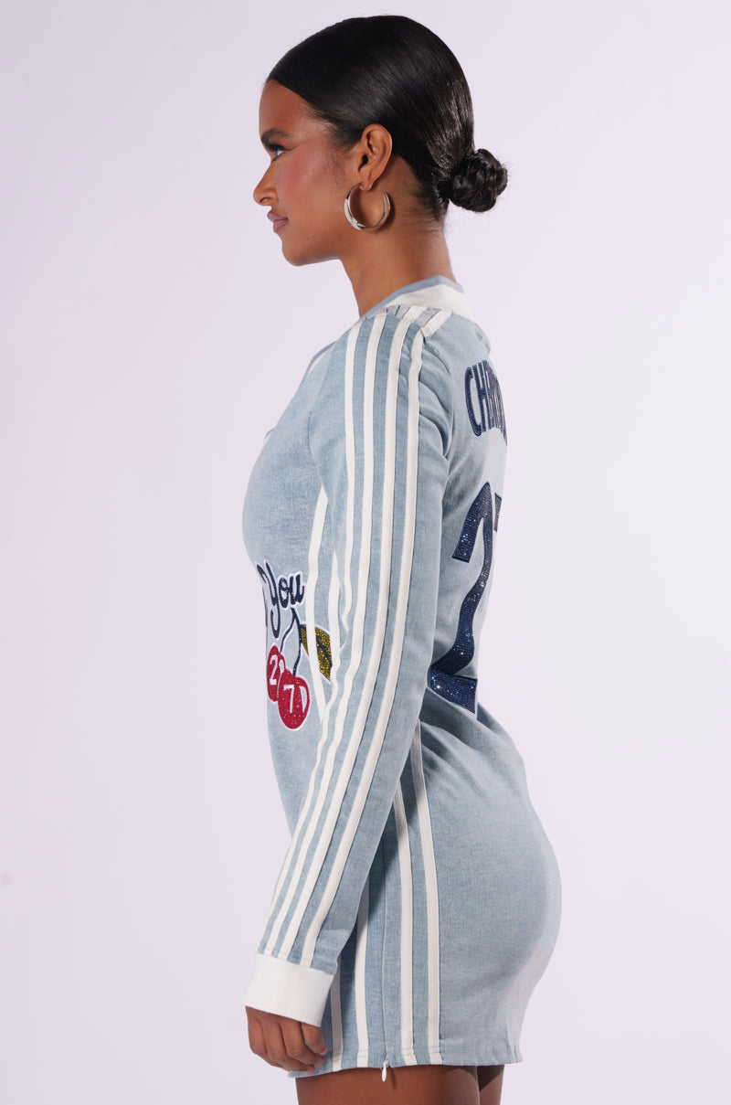 A person with hoop earrings and a sleek bun is wearing the "WITH A CHERRY ON TOP DENIM MINI DRESS," a long-sleeved, light blue dress featuring white stripes on the sleeves. Adorned with CHERRYBOMB 