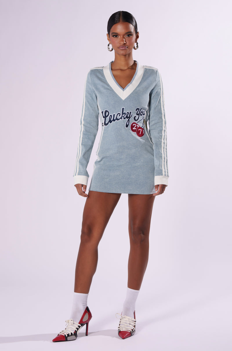 A woman stands facing the camera wearing the "WITH A CHERRY ON TOP DENIM MINI DRESS," a light blue long-sleeve dress featuring white stripes on the sleeves and a "Lucky You" graphic on the front. She pairs the dress with white socks and red and white heeled shoes. She has a neutral expression and her hair is pulled back.
