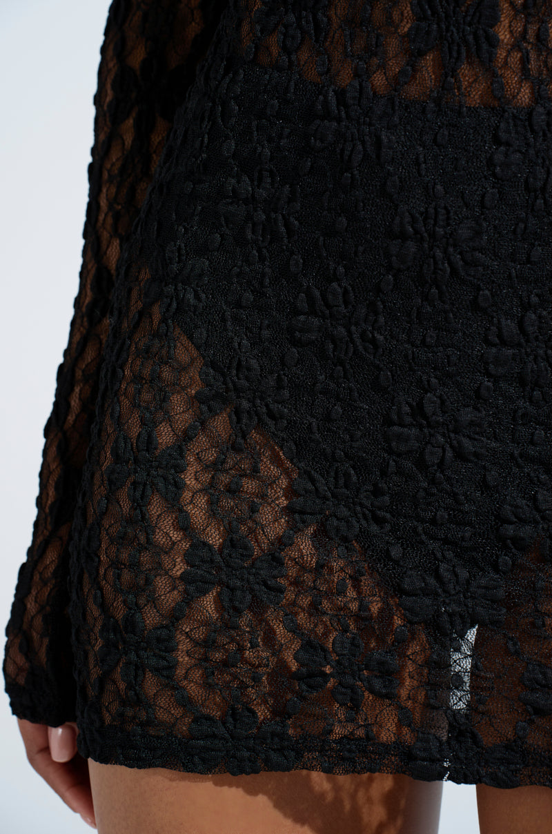 Close-up of a person wearing the LIZZIE LACE MINI DRESS IN BLACK, featuring sheer black lace with a floral pattern. The curve-hugging mini dress subtly reveals skin beneath its see-through texture, highlighting their hand and thigh.
