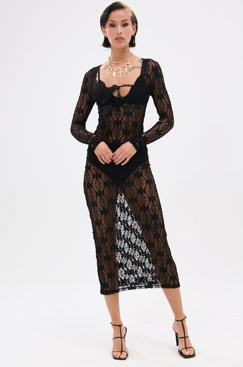A woman with short dark hair poses confidently against a white background in the LIZZIE LACE MAXI DRESS—a sheer mesh maxi with a floral lace base—styled with black undergarments, strappy heels, and layered gold necklaces.