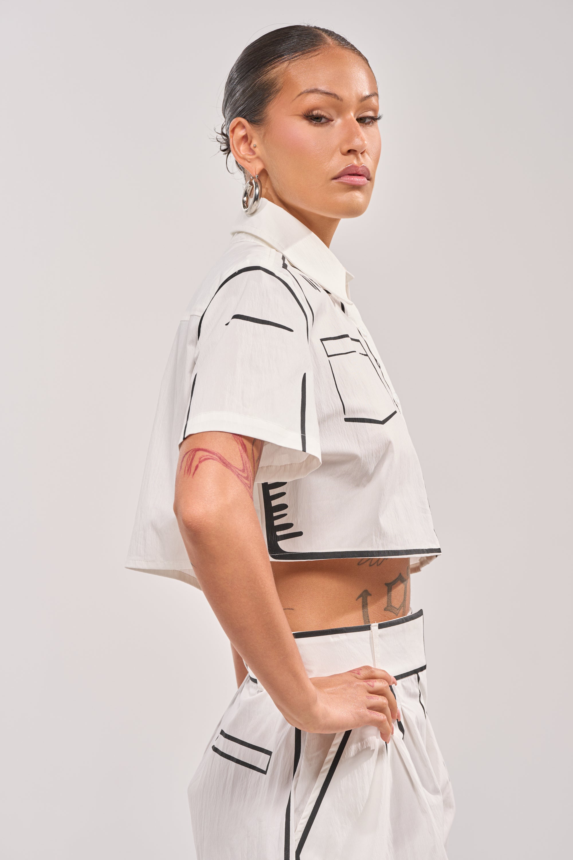 A person with slicked-back hair poses confidently in the BLANK SPACE CROPPED SHORT SLEEVE BUTTON DOWN, a pop-art inspired shirt with bold black outlines and geometric designs, showcasing tattoos on their arm and waist. 1