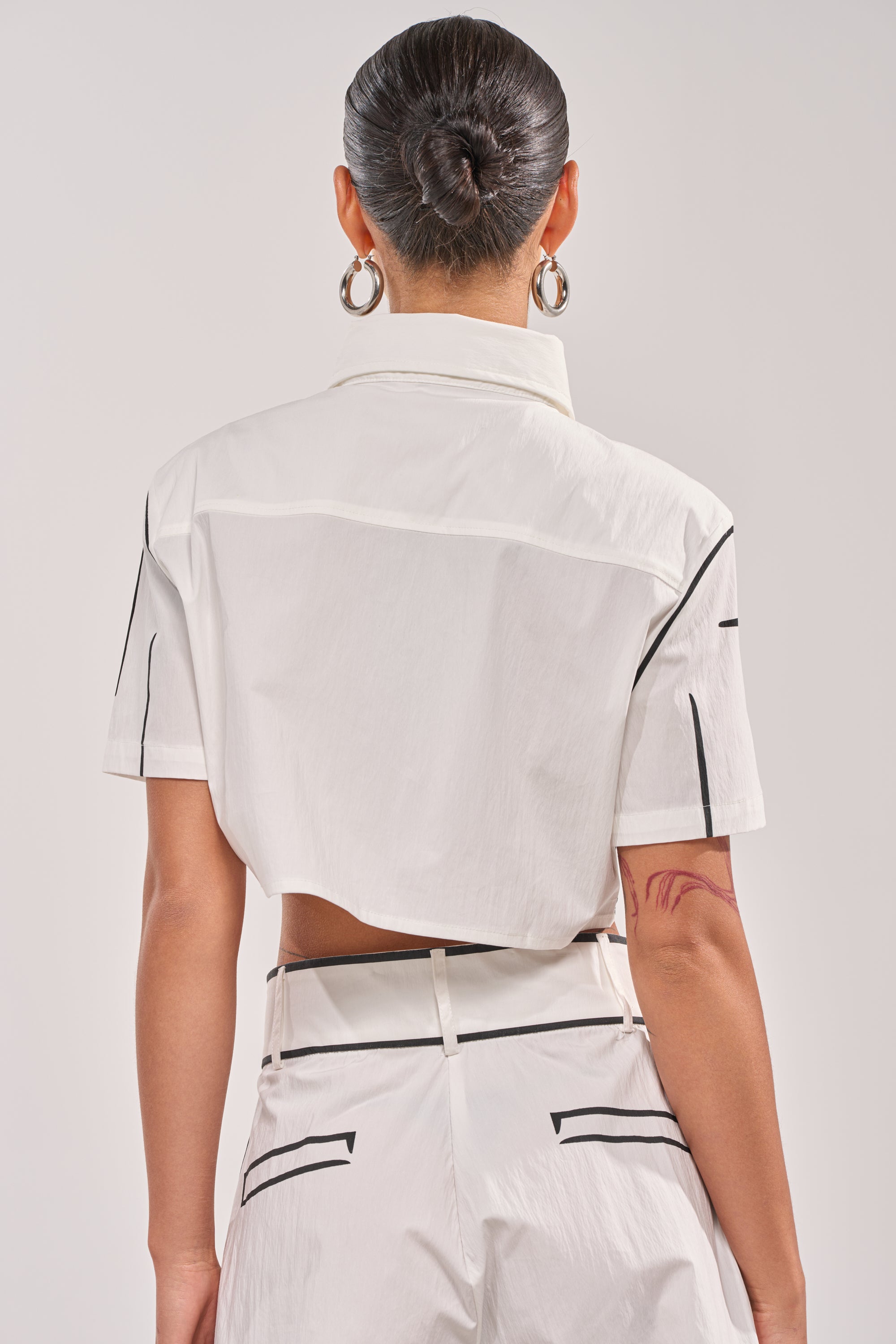 A person with a neat bun and hoop earrings is shown from the back, wearing the BLANK SPACE CROPPED SHORT SLEEVE BUTTON DOWN in white cotton-blend with matching high-waisted pants featuring black trim accents. 2