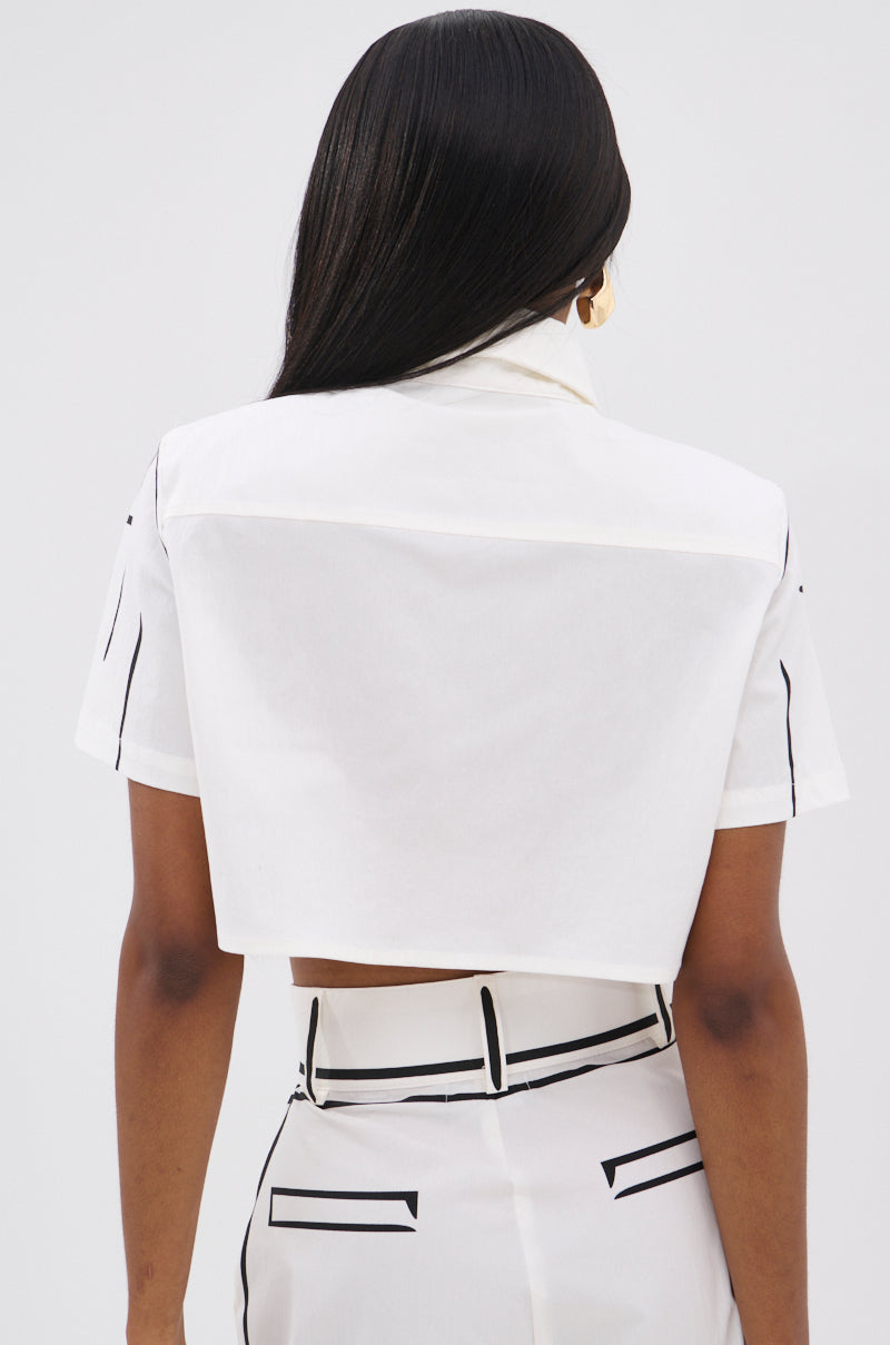 A woman with long, straight black hair is shown from the back in the BLANK SPACE CROPPED SHORT SLEEVE BUTTON DOWN and high-waisted white pants with black accents, standing against a light gray background.