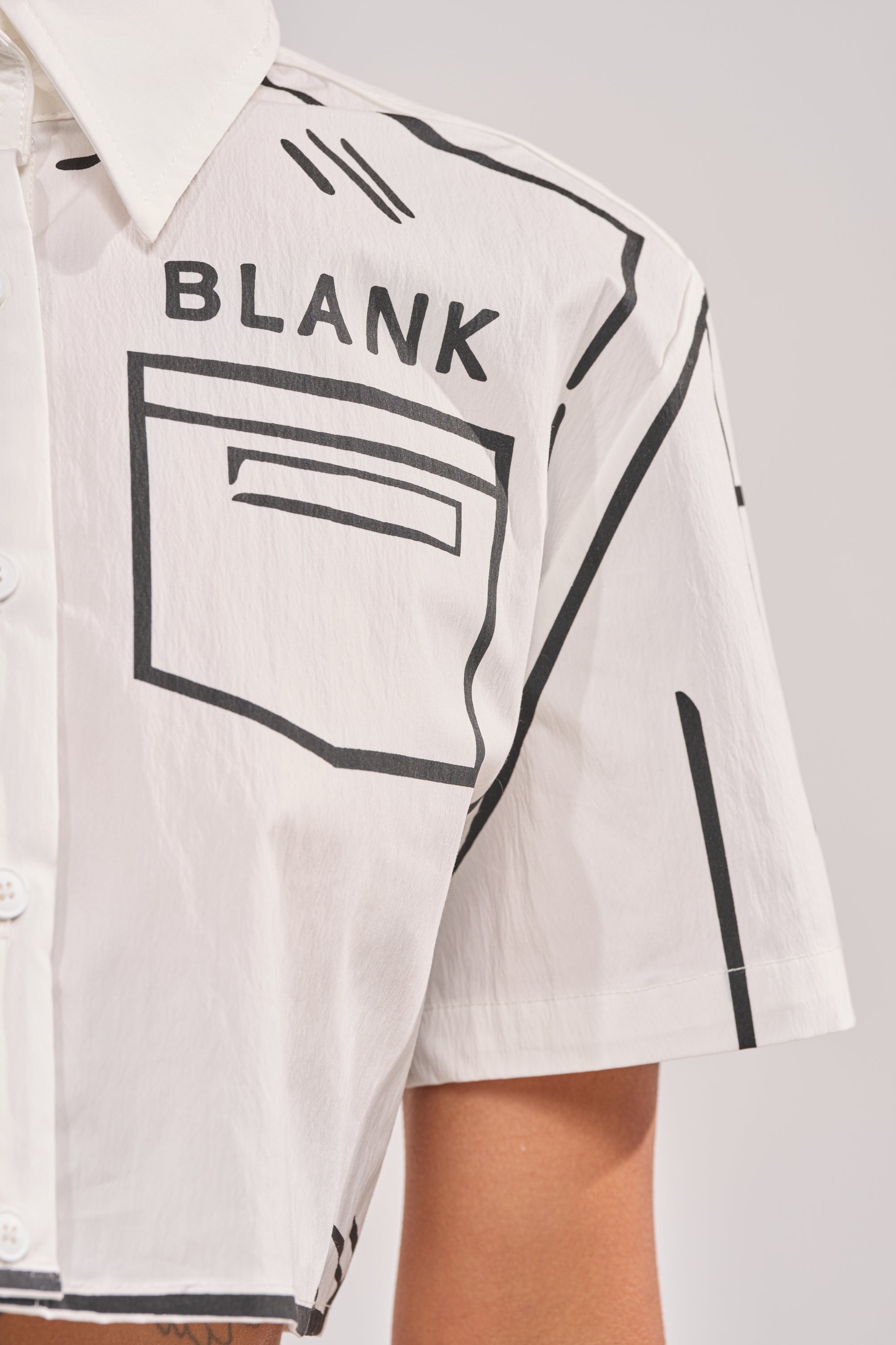 A close-up of the BLANK SPACE CROPPED SHORT SLEEVE BUTTON DOWN: a white cropped shirt featuring black graphic lines and "BLANK" printed above a sketched pocket design on the chest. 5
