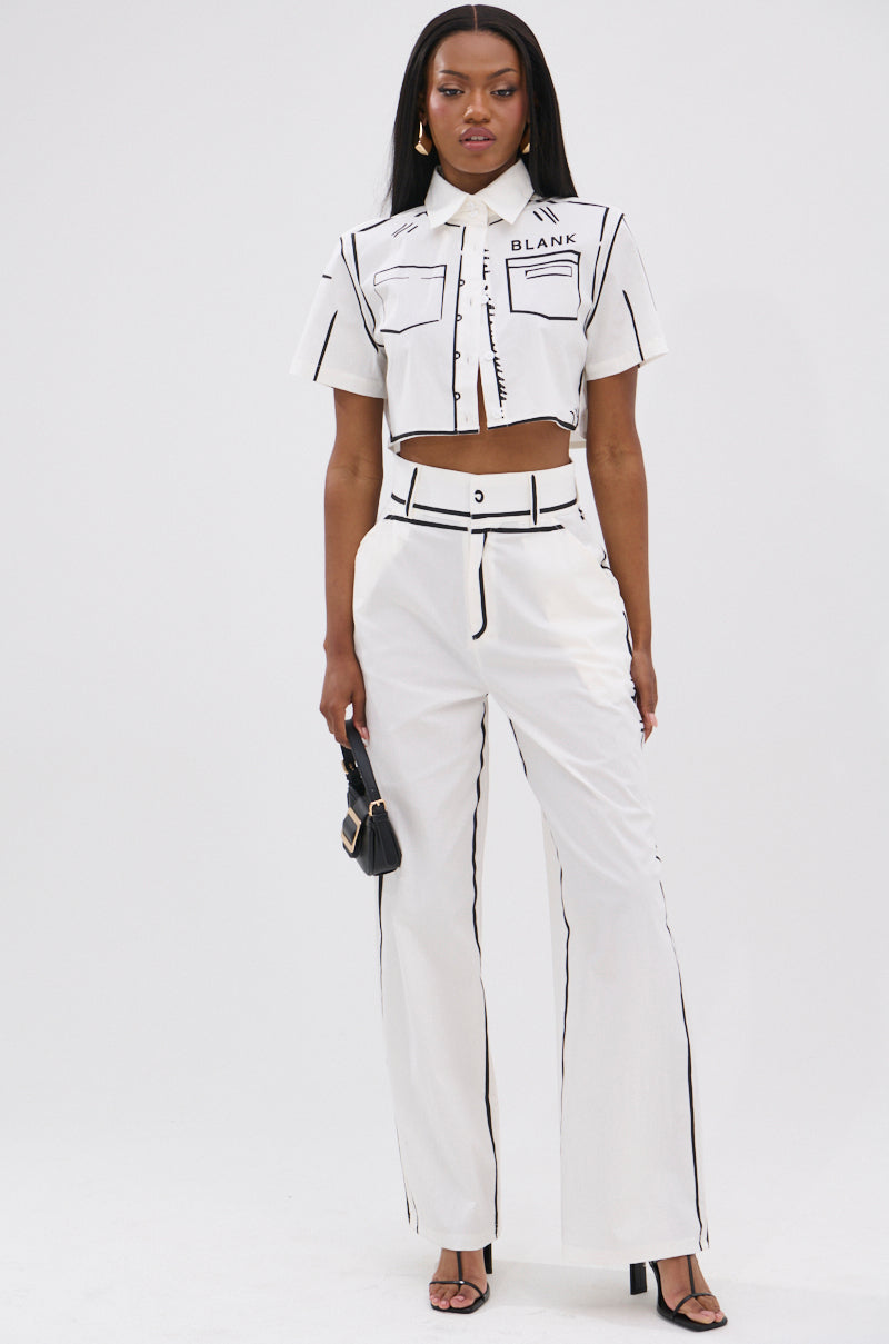 A woman stands against a white background, wearing the BLANK SPACE CROPPED SHORT SLEEVE BUTTON DOWN with matching high-waisted pants. She holds a small black handbag and wears black heeled sandals.