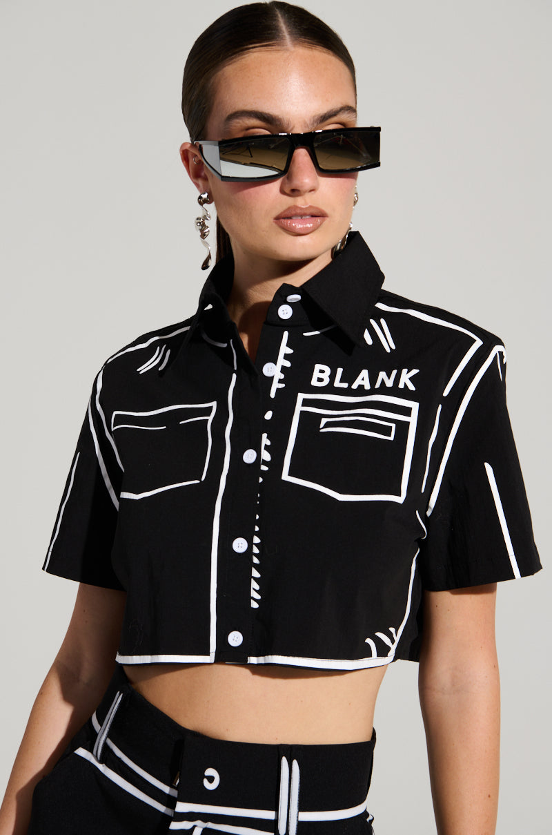 A woman with slicked-back hair is donning large, angular sunglasses and the BLANK SPACE BUTTON DOWN CROP SHIRT from the AKIRA Label—a black, short-sleeve button-up crop top featuring white line detailing and the word "BLANK" on the chest. She pairs it with matching black pants or a skirt while standing against a plain background.