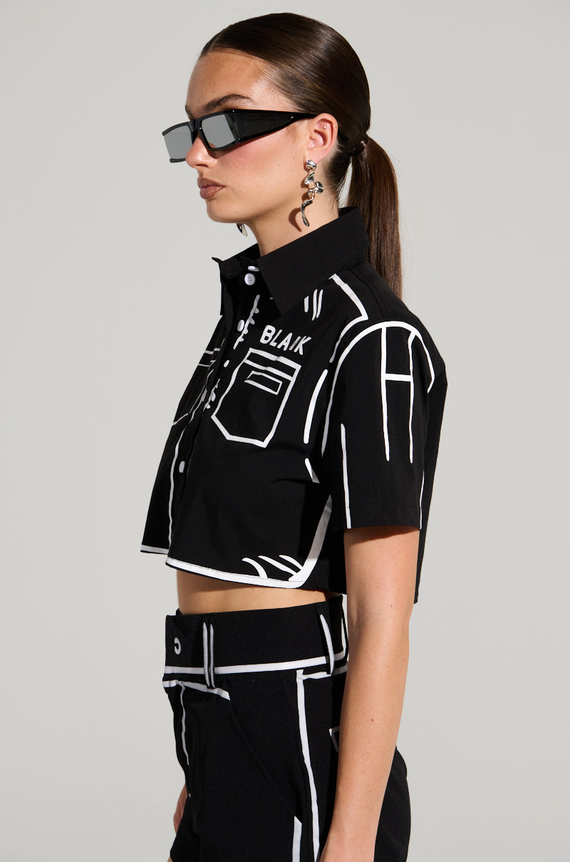 A person stands in profile, showcasing the BLANK SPACE BUTTON DOWN CROP SHIRT from the AKIRA Label with a graphic sketch-inspired print, paired with matching black pants. They wear large, rectangular dark sunglasses and have long hair styled in a sleek ponytail. The background is plain and light gray.