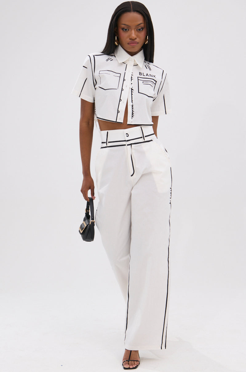 A woman stands confidently in a chic white crop top with black line accents and BLANK SPACE HIGH WAIST PALAZZO PANT—stylish wide-leg bottoms. She accessorizes with a small black handbag and open-toe heels.