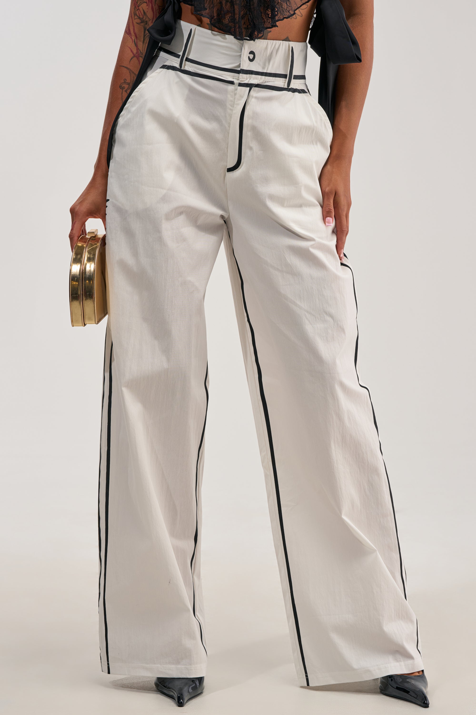 Dressed in the BLANK SPACE HIGH WAIST PALAZZO PANT, this woman showcases crisp white palazzo pants featuring black piping and a sleek wide-leg silhouette. She pairs them with a sheer black lace top and holds a round gold clutch, crafting a striking, statement-ready look ideal for standout concert style. Only her torso and legs are visible.