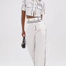 A woman stands confidently in a chic white crop top with black line accents and BLANK SPACE HIGH WAIST PALAZZO PANT—stylish wide-leg bottoms. She accessorizes with a small black handbag and open-toe heels.