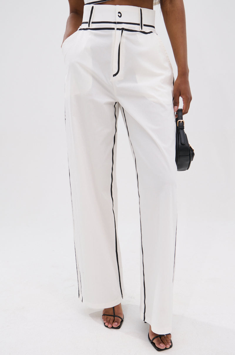 A person wears the BLANK SPACE HIGH WAIST PALAZZO PANT with black piping, black heeled sandals, and a small black handbag against a white background, showcasing chic minimalist style and wide-leg silhouette.