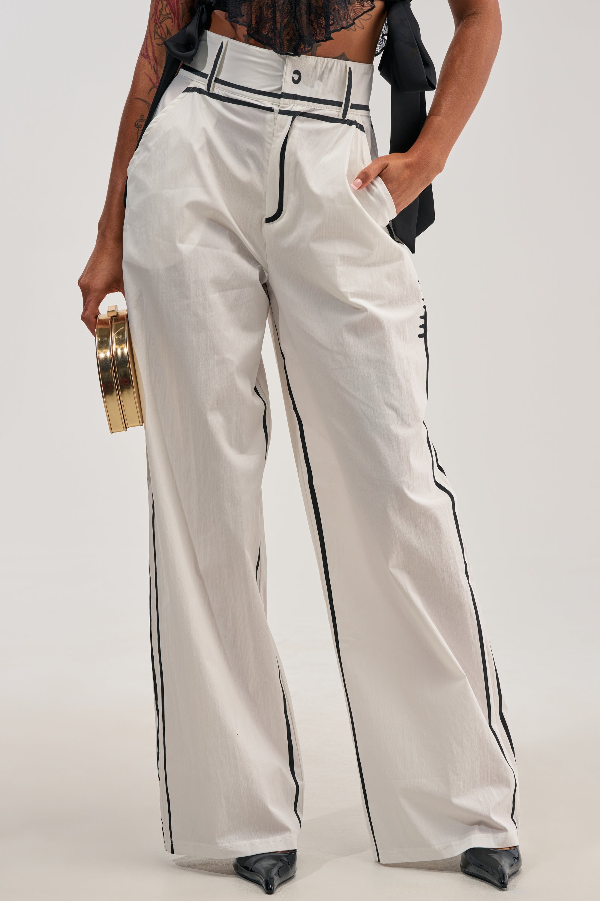 Model wears the BLANK SPACE HIGH WAIST PALAZZO PANT in white, featuring bold black piping details. The wide-leg silhouette stands out, paired with pointed black heels and a round gold clutch—an eye-catching look ideal for making a statement at your next concert. Only the lower half of the ensemble is shown, highlighting the dramatic flair of these palazzo pants.