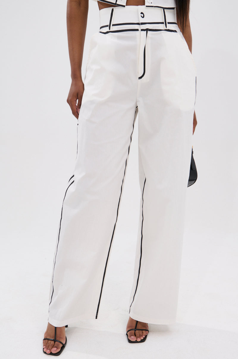 A person models the BLANK SPACE HIGH WAIST PALAZZO PANT with black piping, paired with a crop top and black open-toe heeled sandals, highlighting minimalist style against a plain white background.