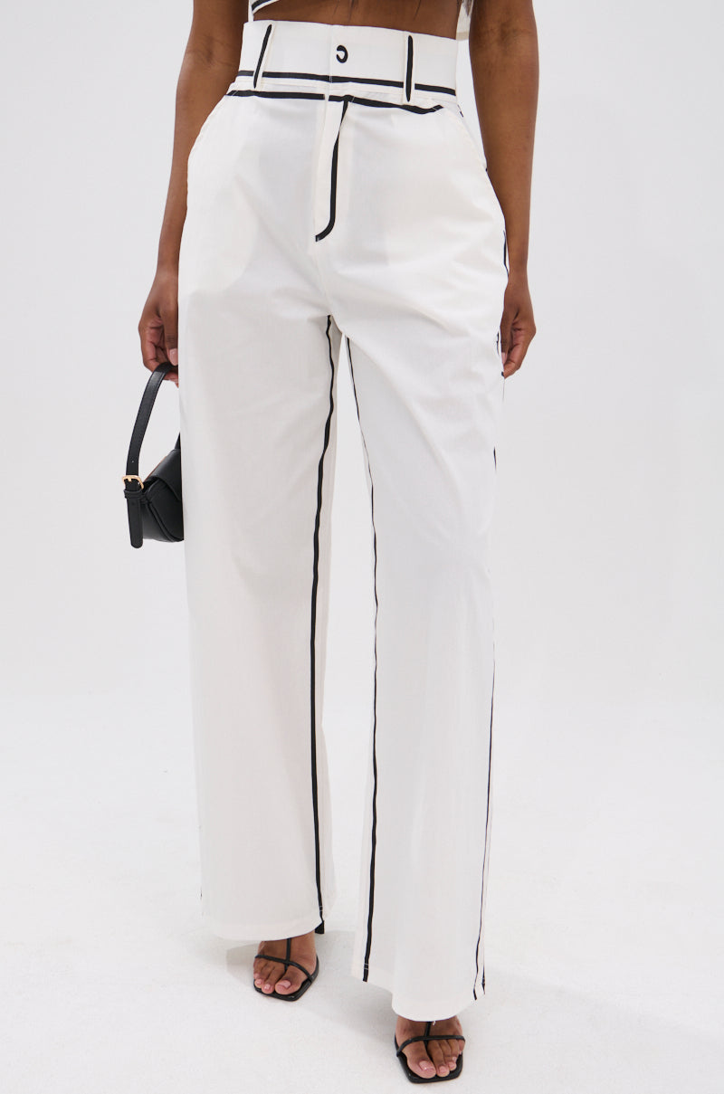 A woman models the BLANK SPACE HIGH WAIST PALAZZO PANT with black piping, styled with a small black handbag and open-toe heels, set against a plain white background for an elegant minimalist look.