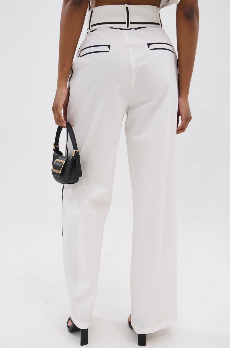 A person stands facing away, wearing BLANK SPACE HIGH WAIST PALAZZO PANT in white with black trim, paired with black open-toe heels and a small black handbag against a plain white background.