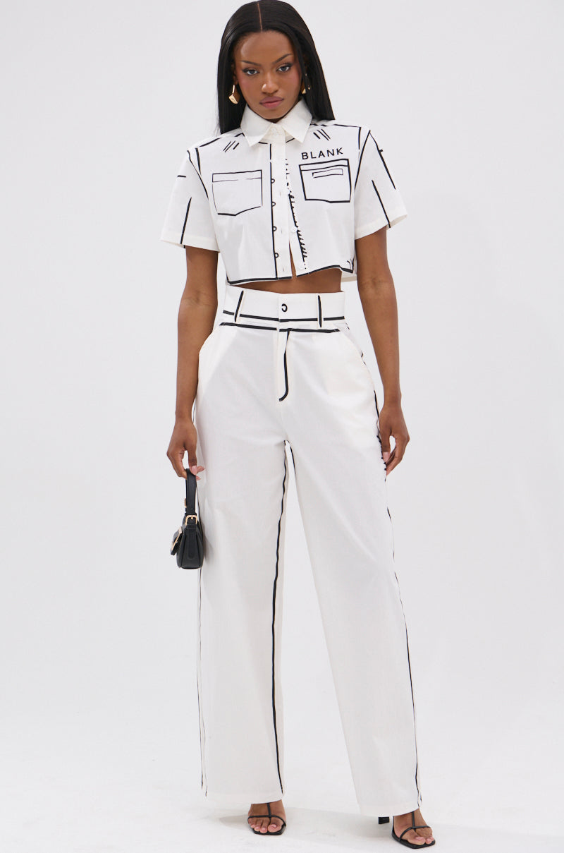 A woman stands against a plain white background, wearing a white short-sleeve crop top and the BLANK SPACE HIGH WAIST PALAZZO PANT with black line details, paired with a small black handbag and black heeled sandals.