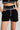 A person is wearing the BLANK SPACE HIGH WAIST FASHION SHORT from the AKIRA Label, which features high-waisted black design with white piping details along the waistband, pockets, and front seam. They are also wearing a matching black cropped top adorned with white accents while keeping their hands in the shorts' pockets.