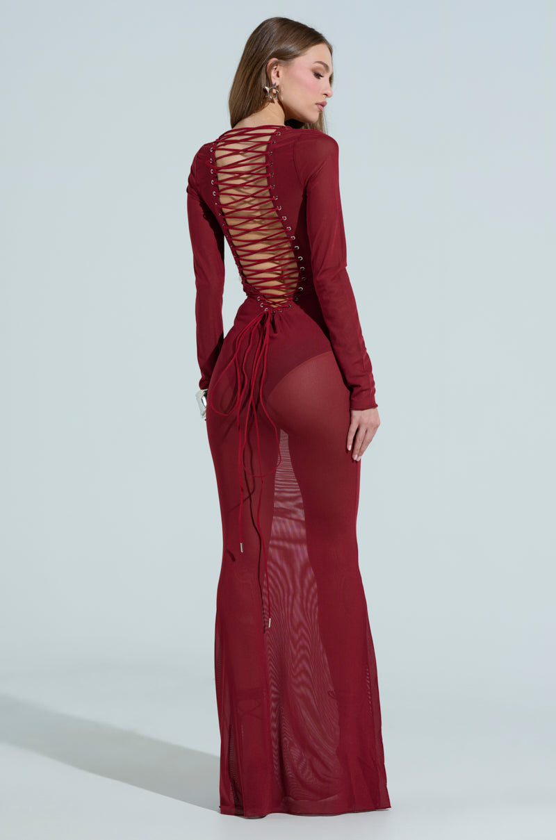 Against a light gray backdrop, a woman showcases her long hair and earrings in the PRIME ENERGY MESH MAXI DRESS. The sheer red gown features a lace-up back and scoop neckline, while its fitted silhouette enhances her sophisticated allure.