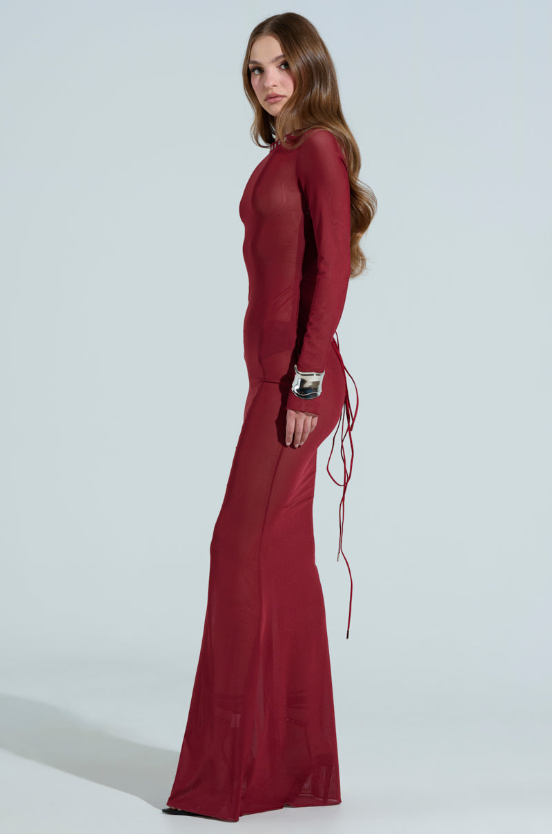 A person with long hair poses in the PRIME ENERGY MESH MAXI DRESS, a form-fitting sheer maroon maxi dress featuring a scoop neckline, lace-up back, and long sleeves against a plain, light-colored background.