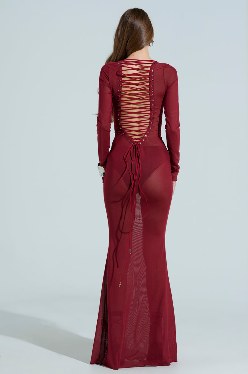 A woman, with long hair flowing loosely, is turned away from the camera while wearing the floor-length PRIME ENERGY MESH MAXI DRESS. This burgundy dress features a lace-up back and a fitted design against a plain background.