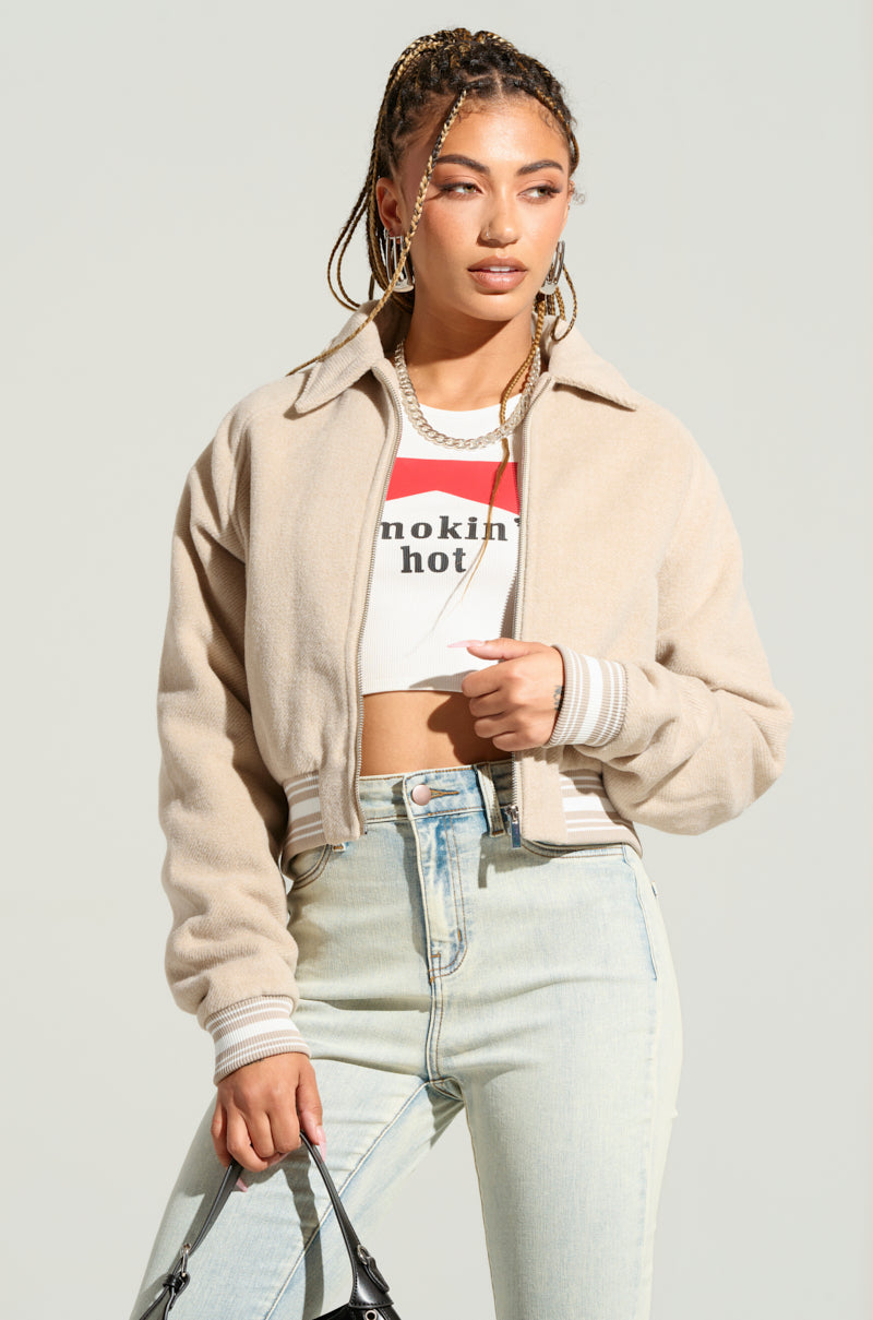 A woman with braided hair sports the LIA WOVEN BOMBER in beige, along with a cropped graphic t-shirt and light denim jeans. She carries a small black purse and stands against a light background, accessorized with hoop earrings and a necklace, exuding effortless style.