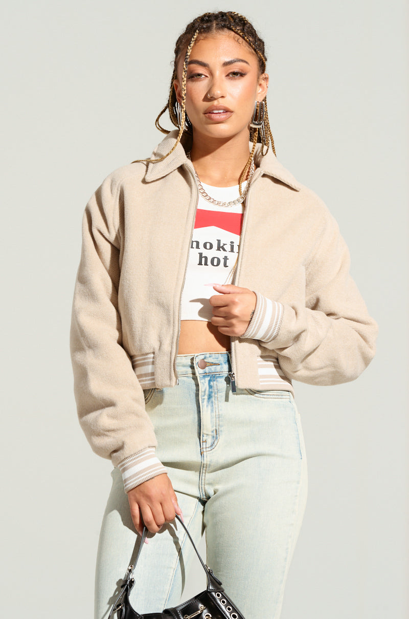 A person with braided hair is wearing the LIA WOVEN BOMBER, which features a quilted interior lining. They pair it with a white text-emblazoned crop top, light blue jeans, and hold a black handbag while standing against a neutral background.